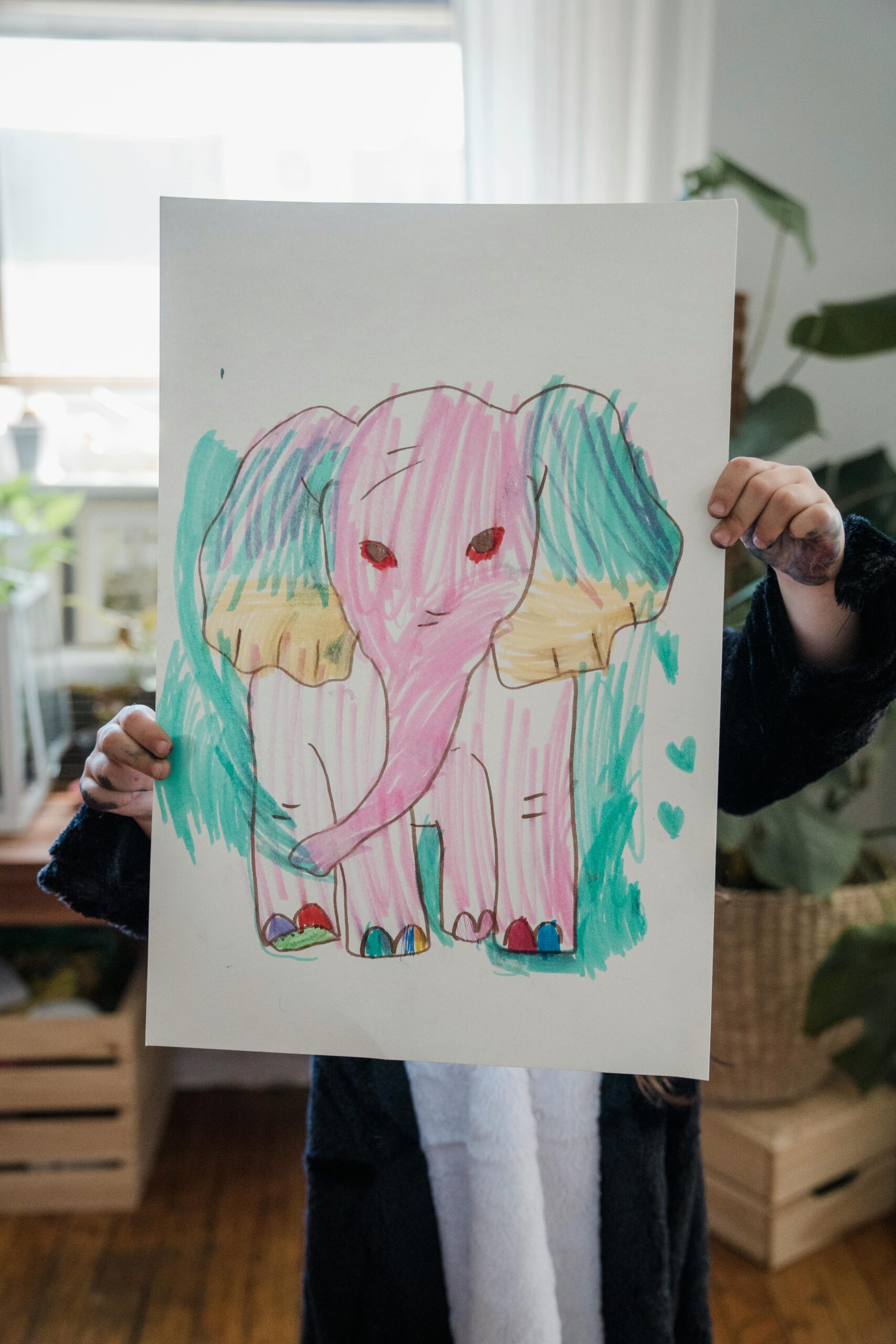 A child proudly displays a colorful elephant drawing indoors, showcasing creativity and art.